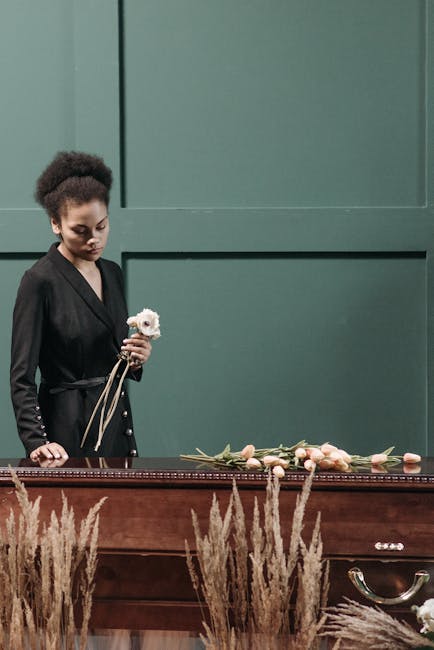 A woman with dark curly hair, dressed in a black blazer with metallic buttons, stands beside a wooden casket at a funeral. She holds a small bouquet of white flowers with green stems. In front of her,