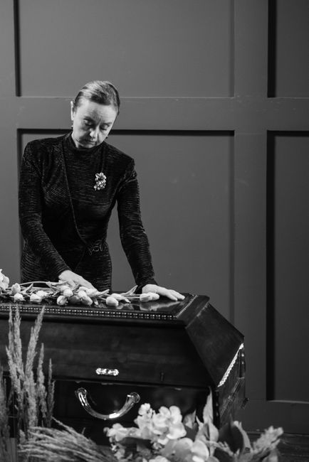 A woman dressed in black, with her hair styled in a neat updo, is arranging a floral tribute on a dark wooden casket during a funeral service. In front of her, there are fresh white and pale pink flow