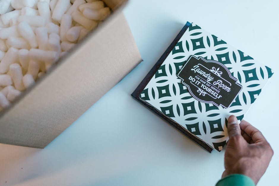 A hand holding a floral gift box with a black and white geometric pattern and a label that reads 'Laundry Gift for Same Day Service, Do It Yourself.' The box is being opened, revealing white packing p