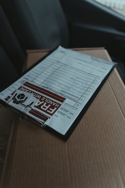 A close-up of a wrapped floral bouquet placed on top of a cardboard box, with a delivery receipt or invoice attached. The bouquet includes a variety of fresh flowers such as pink roses, white lilies,