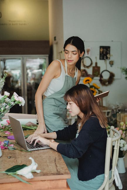 Two women in a flower shop are engaged in floral arrangements or ordering. One woman with long, dark hair wearing a light-colored apron stands beside the table, observing or assisting, while the other