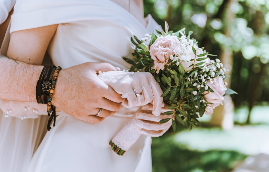 A bride's hands holding a bouquet of fresh, pastel pink and white roses, complemented by delicate white baby's breath and lush green foliage, wrapped in a white lace ribbon. The bride is wearing a whi