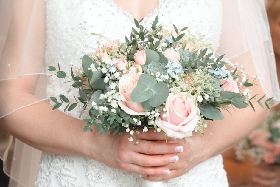 A bride holding a delicate floral bouquet featuring soft pink roses, white baby's breath, and green eucalyptus leaves, arranged in a rounded, compact style. The bouquet is wrapped with elegant white r
